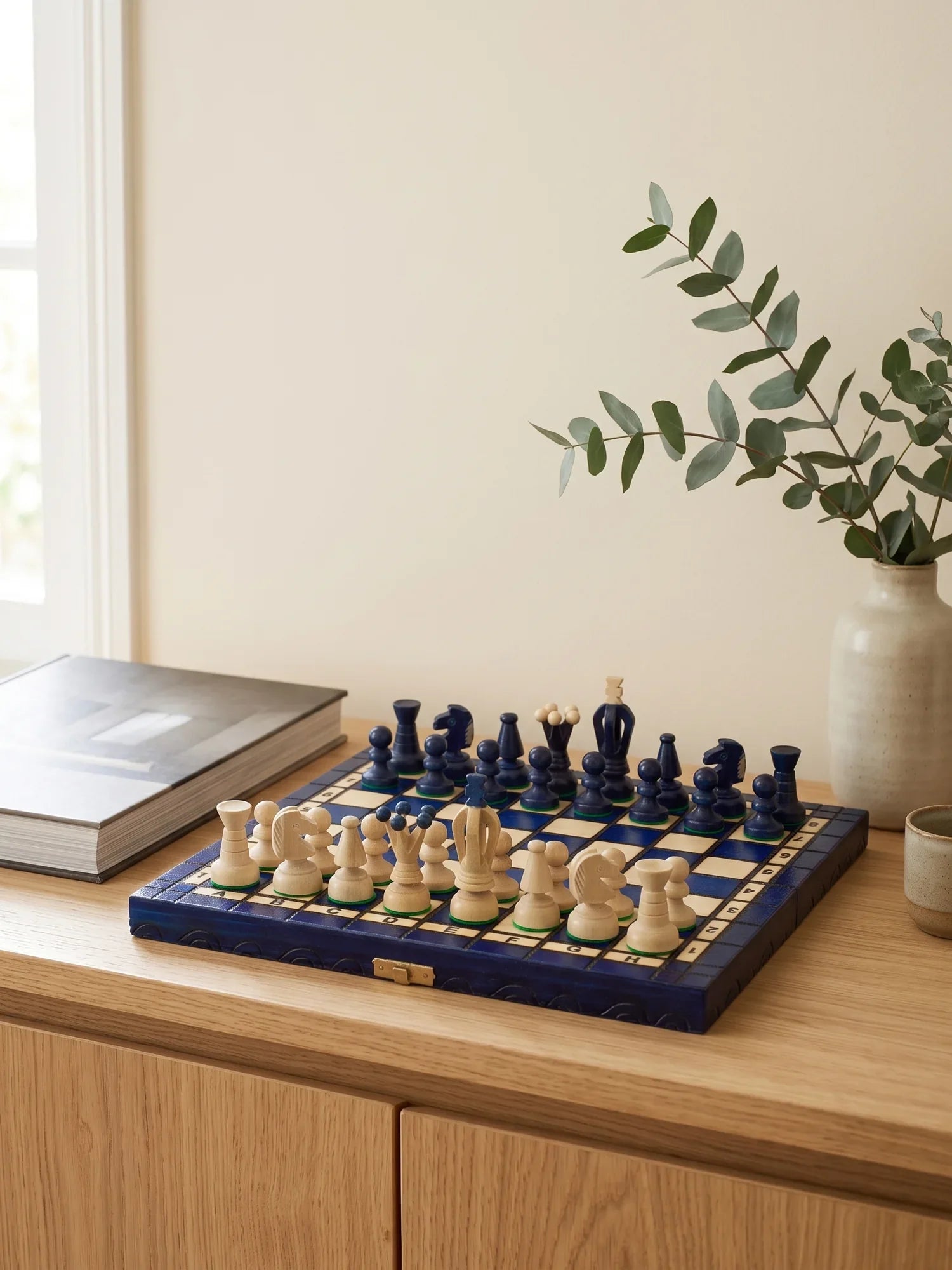 Échiquier pliant bleu posé sur un buffet en bois clair, à côté d’un livre d’art et d’un vase d’eucalyptus